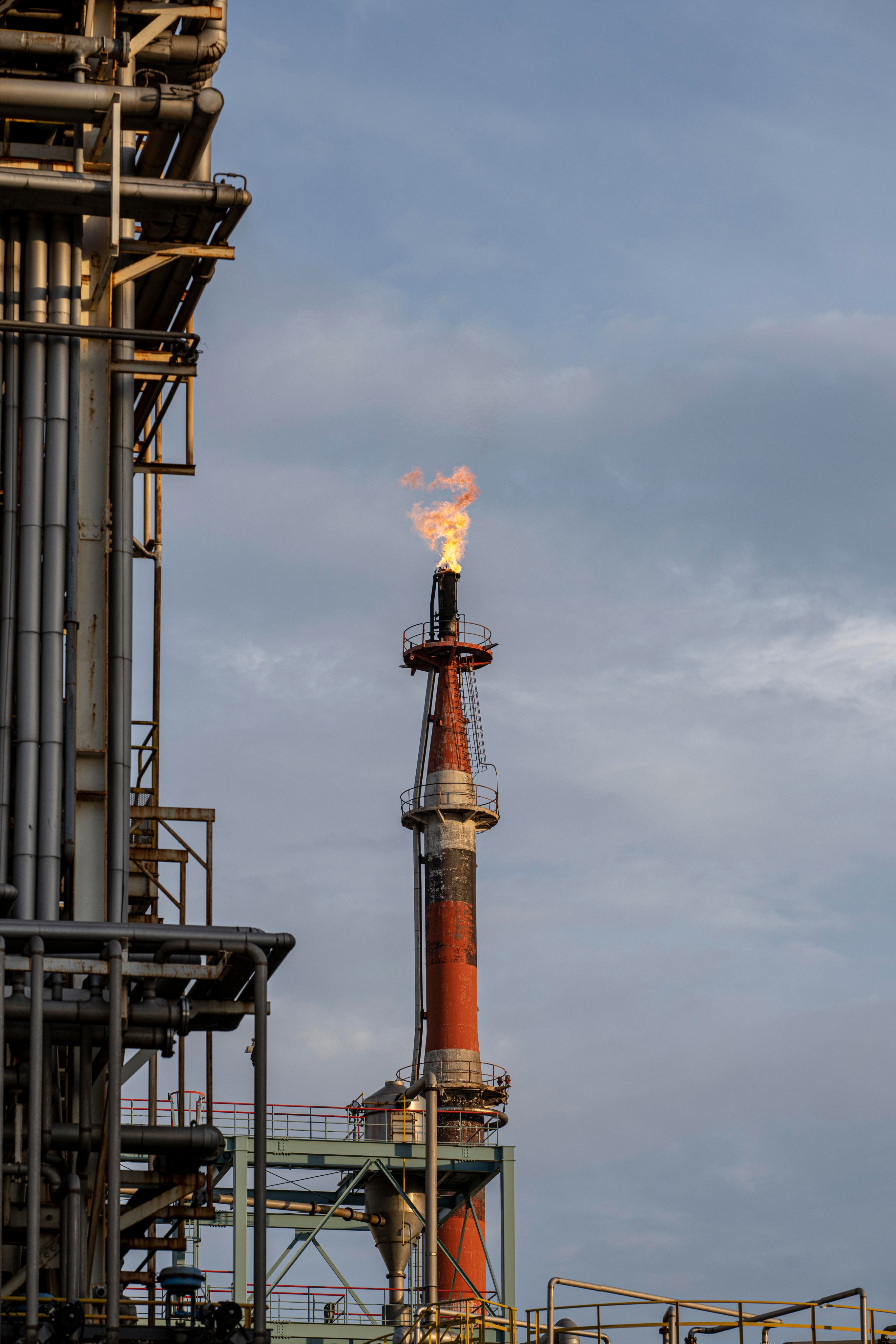 A tall industrial flare stack at a refinery emitting a bright orange flame against a cloudy sky.