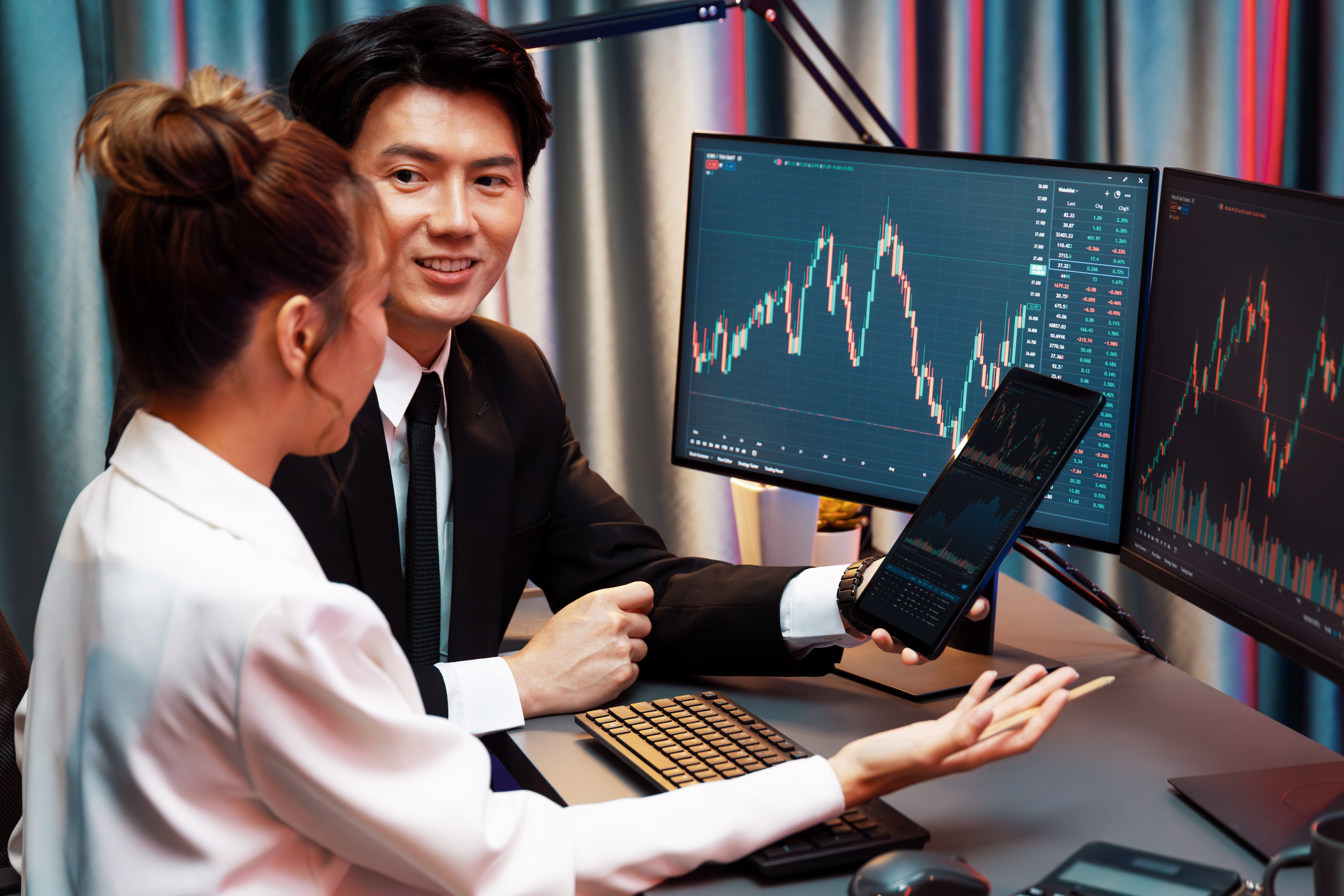 A smiling businessman showing a trading chart on a tablet to a colleague in an office setting with a large monitor displaying financial data.