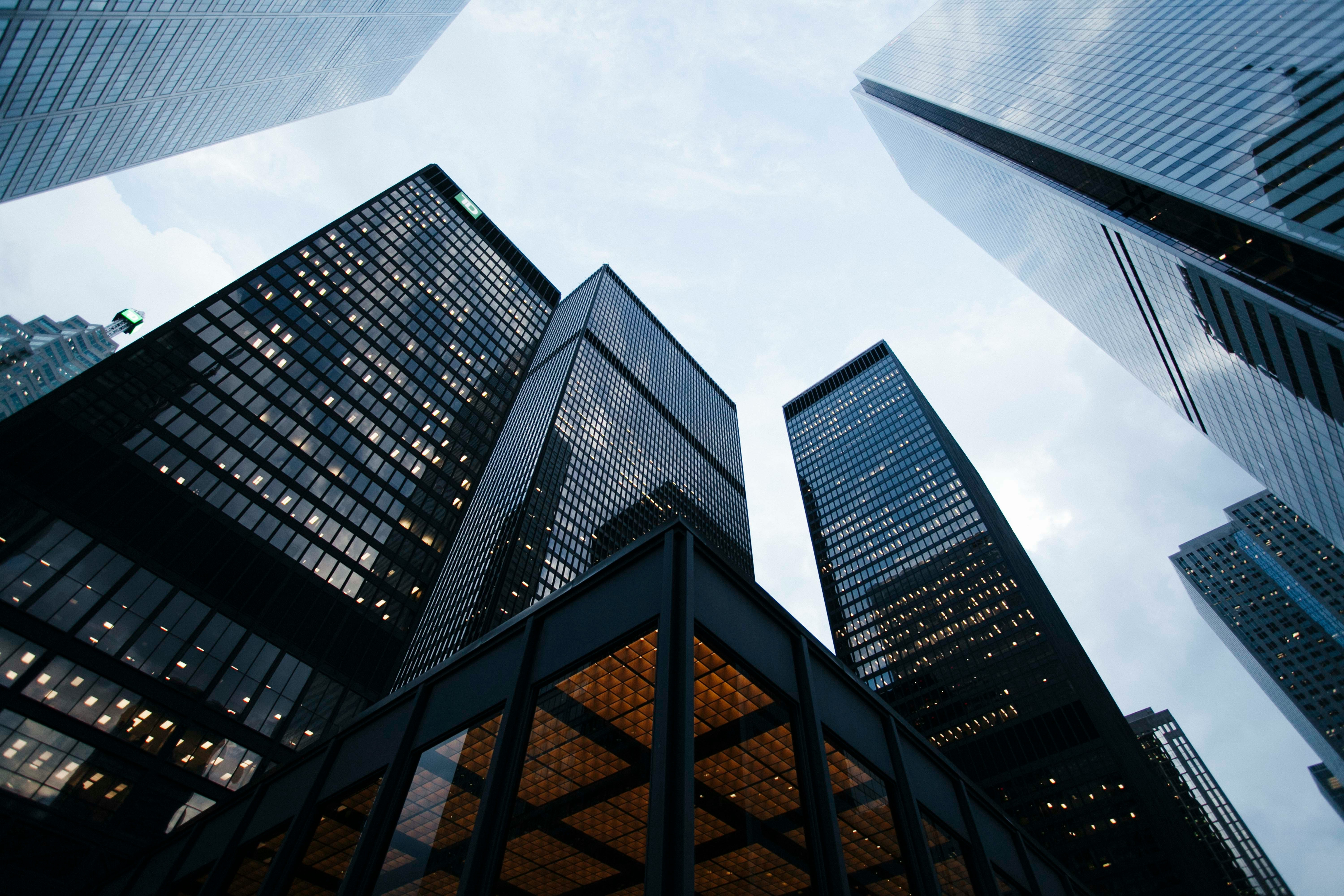 A low-angle view of modern glass skyscrapers reaching toward a cloudy sky in a dense urban financial district.
