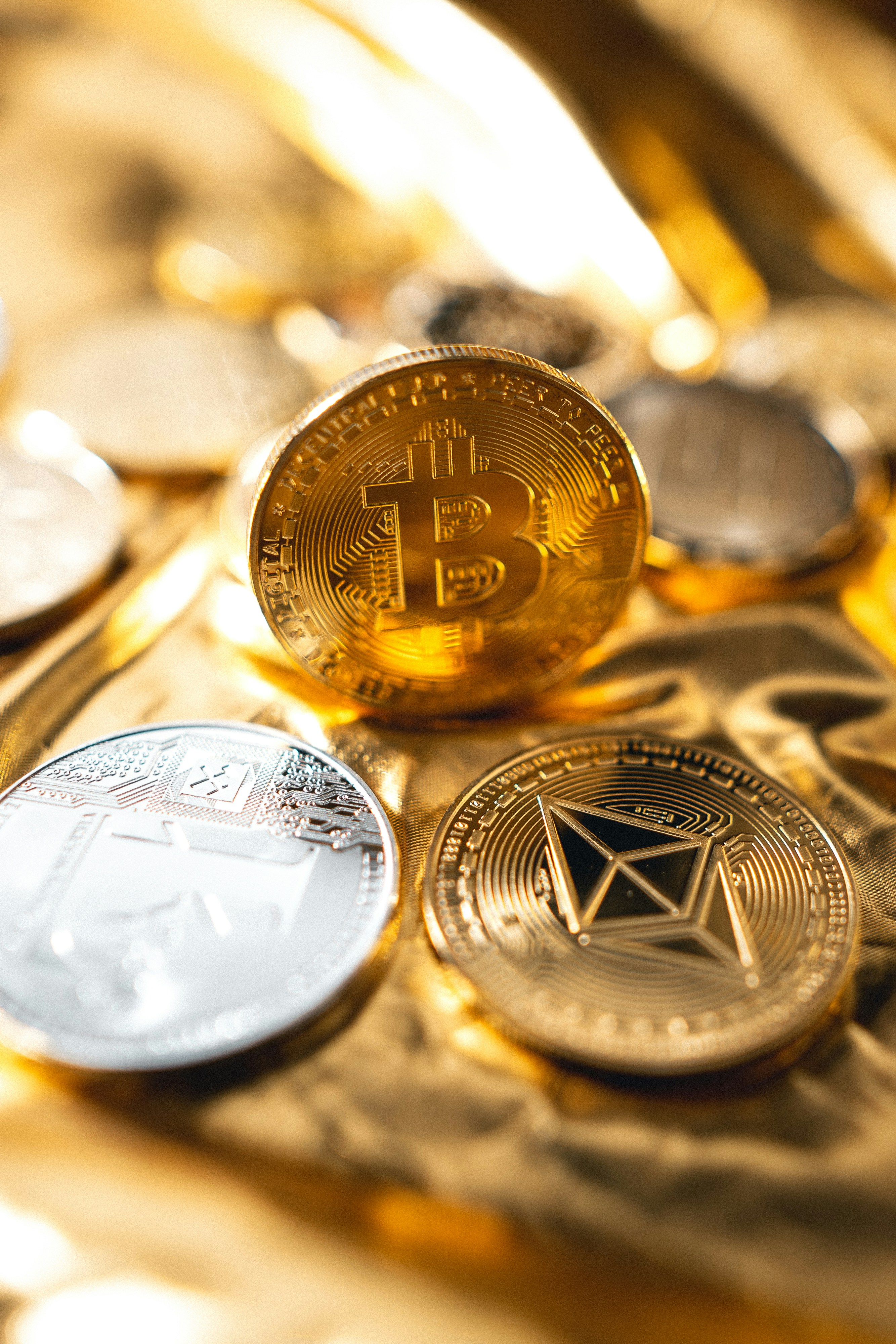 A gold Bitcoin coin stands upright on a shimmering gold surface, flanked by a silver coin and a gold Ethereum coin in the foreground.