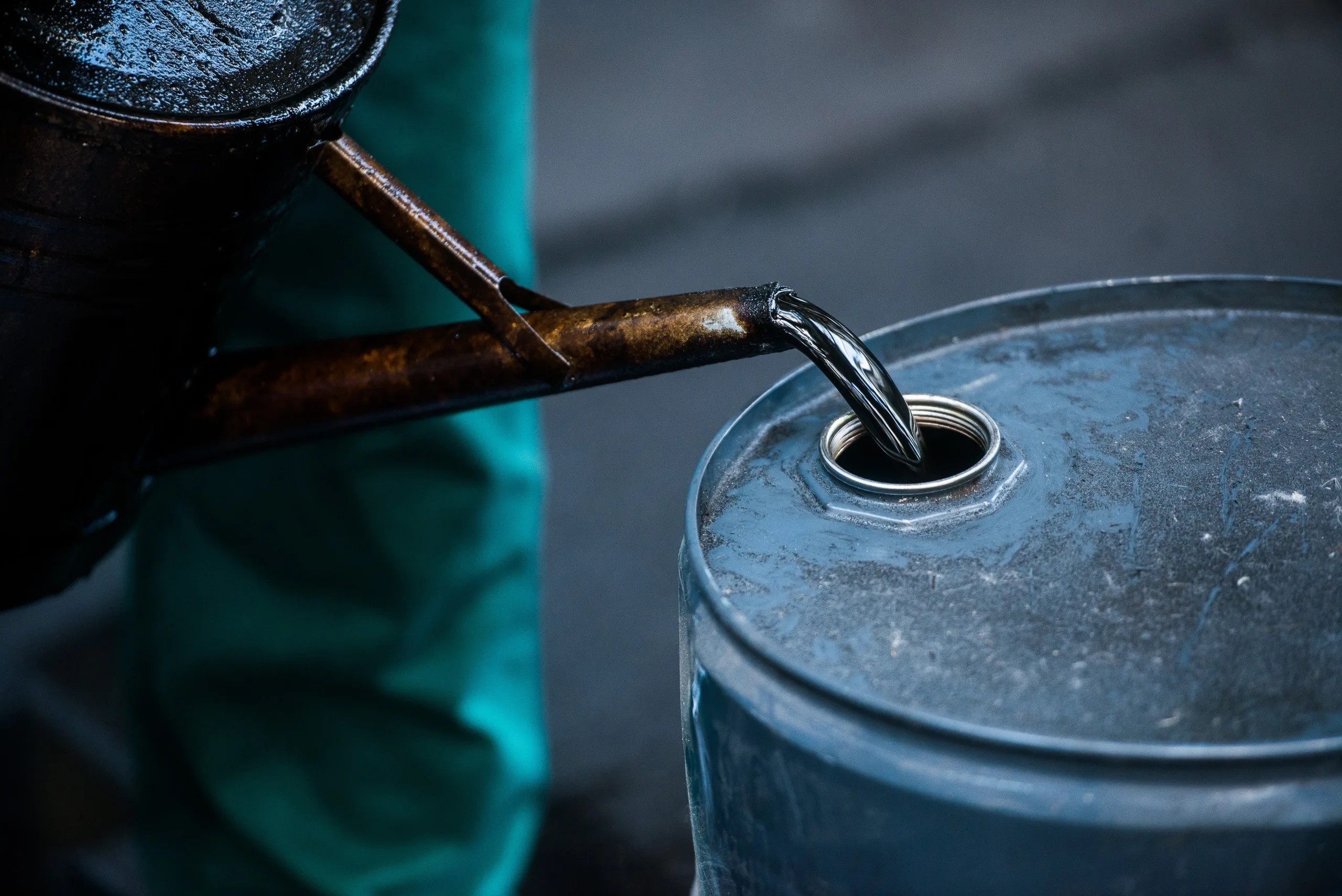 Thick black oil being poured from a spout into a blue metal barrel.
